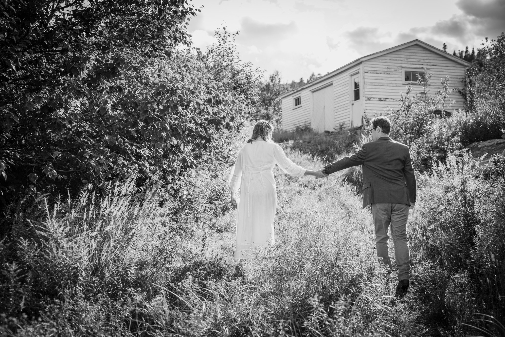Couple walking through wild grass holding hands with old building in the background while smiling at each other.